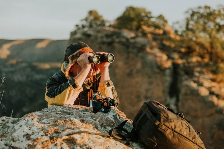A person carrying a camera and using binoculars on a rocky overlook.