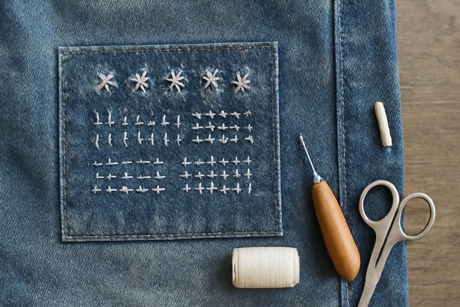 A denim patch with visible mending stitches in white thread forming a sampler of simple patterns